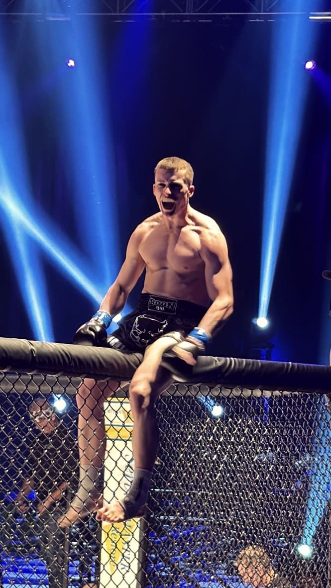 A TCB fighter sits on top of the cage under blue arena lights after a win.