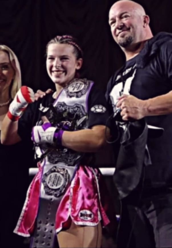 A TCB fighter smiles with her championship belt beside her coach.