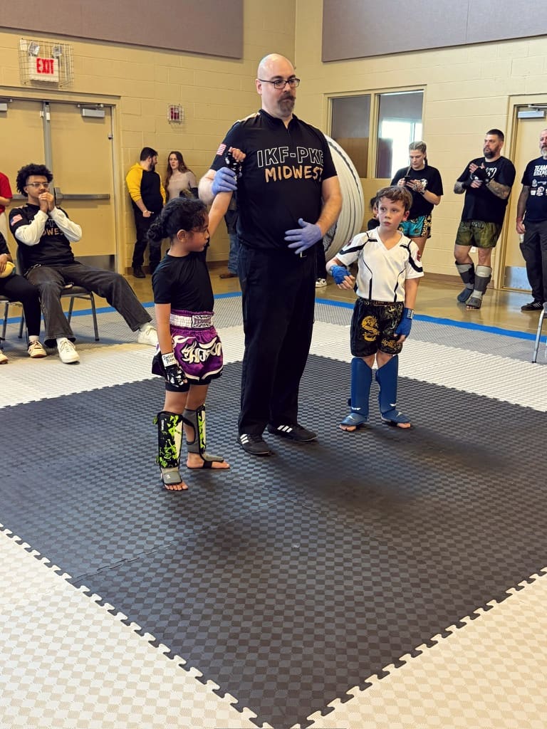Two young fighters in headgear square off on the mat as a referee looks on.