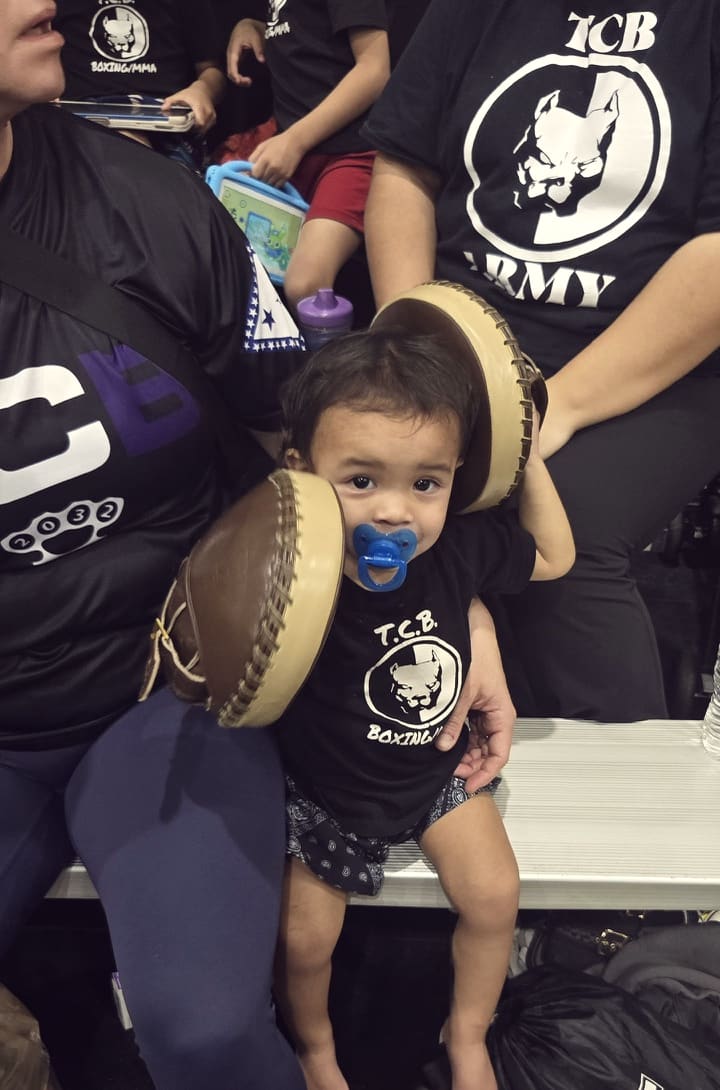 A toddler in a TCB Army shirt holds up a pair of focus mitts.