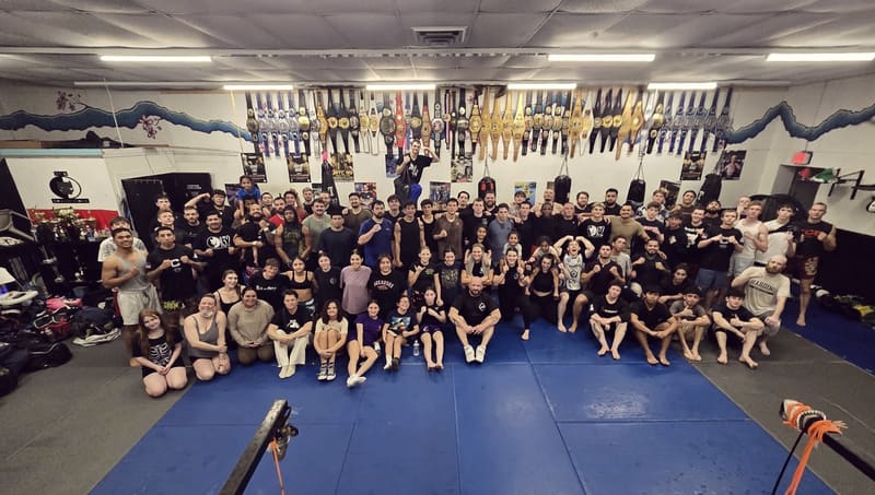 Dozens of TCB members, kids and adults, pack the mats for a team photo under a wall of championship belts.
